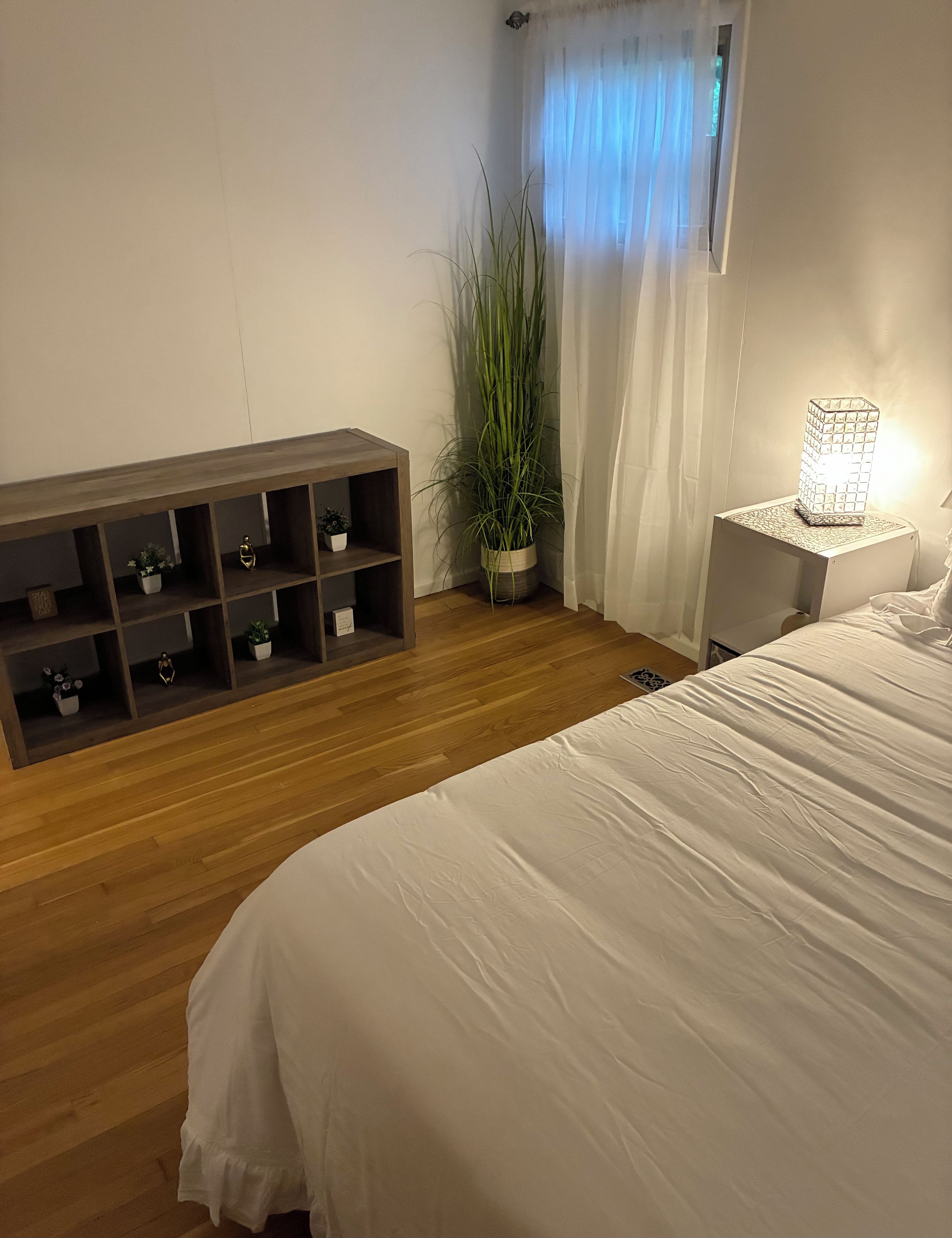 Un dormitorio con piso de madera, una cama con ropa de cama blanca, una mesita de noche con una lámpara, una planta y un estante decorado con objetos ornamentales.