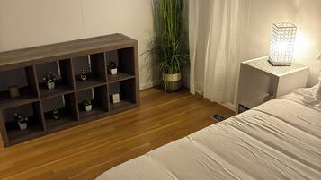 Un dormitorio con piso de madera, una cama con ropa de cama blanca, una mesita de noche con una lámpara, una planta y un estante decorado con objetos ornamentales.