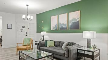 Un salon avec un mur d’accent vert, un canapé gris, une table basse en verre et deux tables d’appoint avec des lampes.