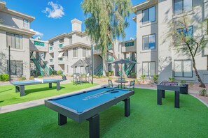 An outdoor pool area with two foosball tables and a pool table, surrounded by a well-maintained lawn and a building with multiple windows and balconies. - Heated Pool | Walk to Westin Kierland - Park-W/D (Scottsdale)
