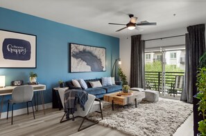 A modern living room with a blue accent wall, a ceiling fan, a large window with curtains, a sofa, a coffee table, and a desk with a chair. - Heated Pool | Workspace | Relaxing Workcation (Phoenix)