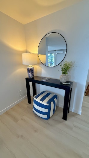 A room with a black console table, a round mirror, a lamp, a small potted plant, and a striped ottoman. - Twin Tides South | Renovated 2BR (Bremerton)