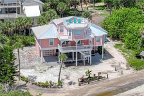 A two-story house with a balcony, surrounded by greenery and a sandy area.