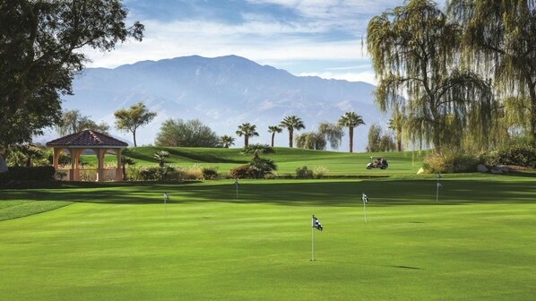Um campo de golfe com um coreto, palmeiras e montanhas ao fundo.