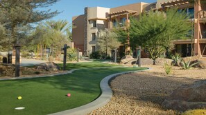 Um campo de golfe com putting green, rodeado por áreas ajardinadas e um edifício de vários andares com varandas.