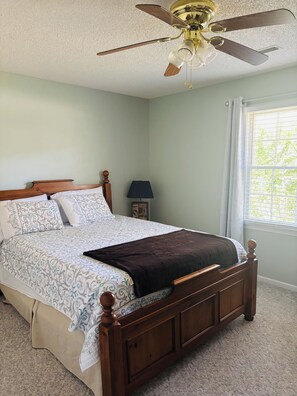 A bedroom with a wooden bed, a ceiling fan, a nightstand with a lamp, and a window with curtains. - Amazing 3-bedroom house with private lake access and dock, (Manning)