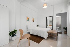 A modern, minimalist bedroom with a bed, bedside table, chair, and two potted plants. - San Rafael Apt 8 | Elegant 1 BR Stay @ San Juan (San Juan de Puerto Rico)