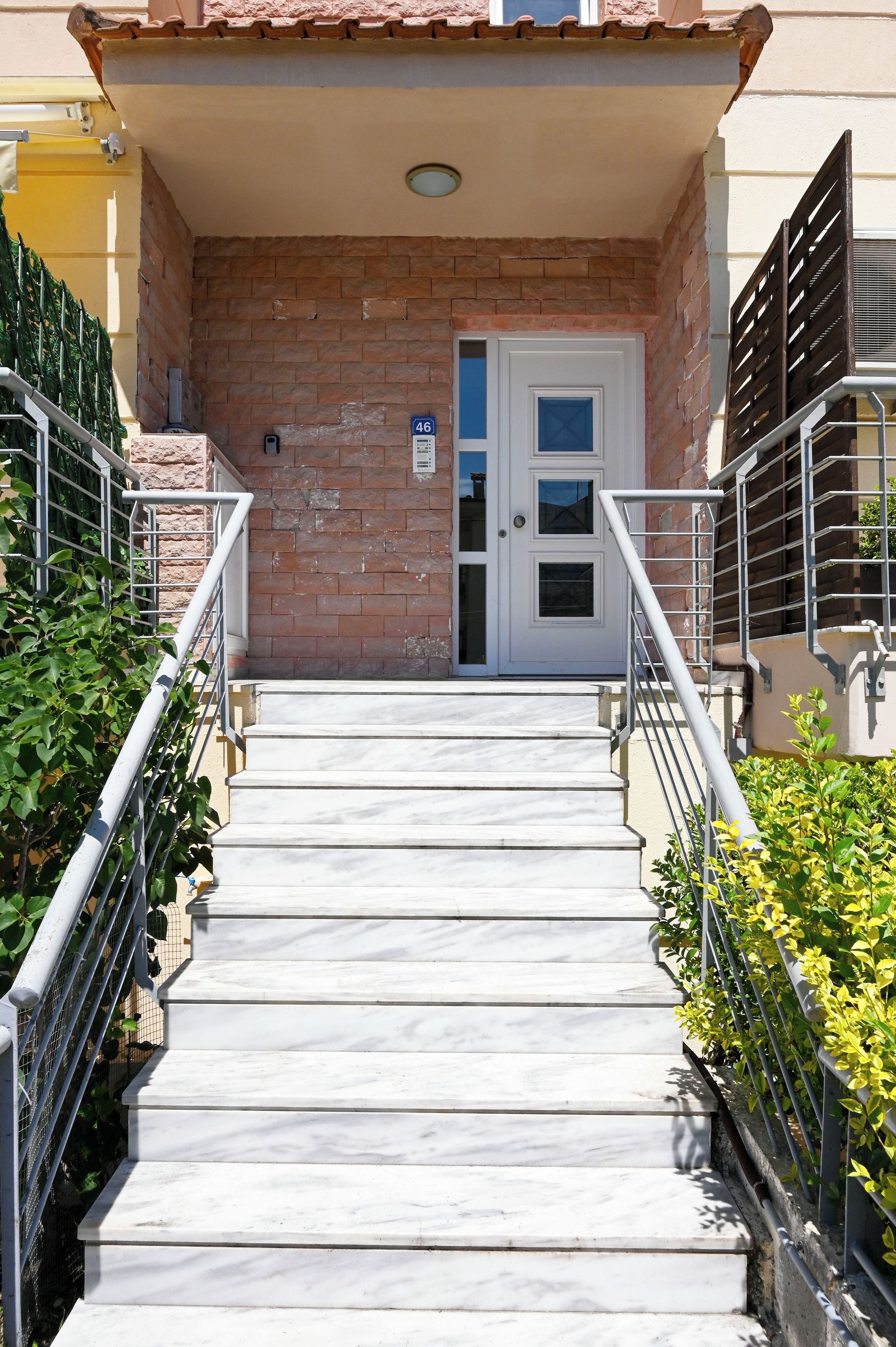 A set of marble stairs leading to a white door with a blue window, flanked by metal railings and greenery.