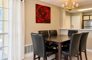 A dining area with a table and chairs, a chandelier, a red rose painting, and a kitchen in the background. - ATX Basecamp - Near The Domain & Q2 Stadium (Austin)
