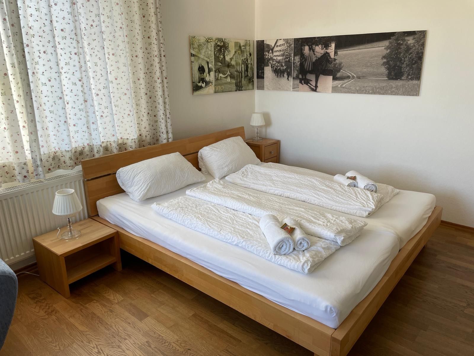 Un lit bien fait, avec des draps blancs et des serviettes pliées, une tête de lit en bois, une table de chevet avec une lampe et un mur orné de photographies encadrées.