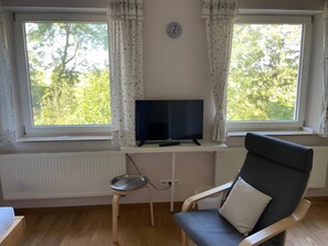 A room with a television on a stand, a chair with a cushion, a small table, and a window with curtains. - Studio Apartment 'Marleen' with Wi-Fi (Ottobeuren)