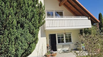 Ein zweistöckiges Haus mit Balkon, Eingangstür und kleiner Terrasse mit Gartenmöbeln.