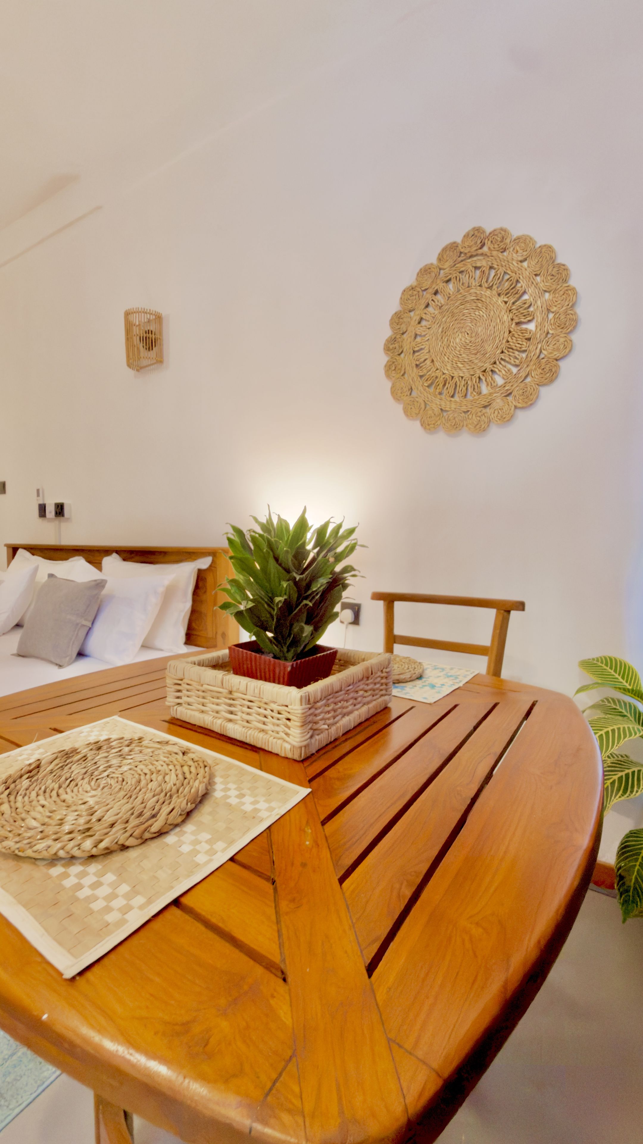 Une table de salle à manger en bois, agrémentée de sets de table tressés, d’une plante en pot et d’un élément décoratif fixé au mur.