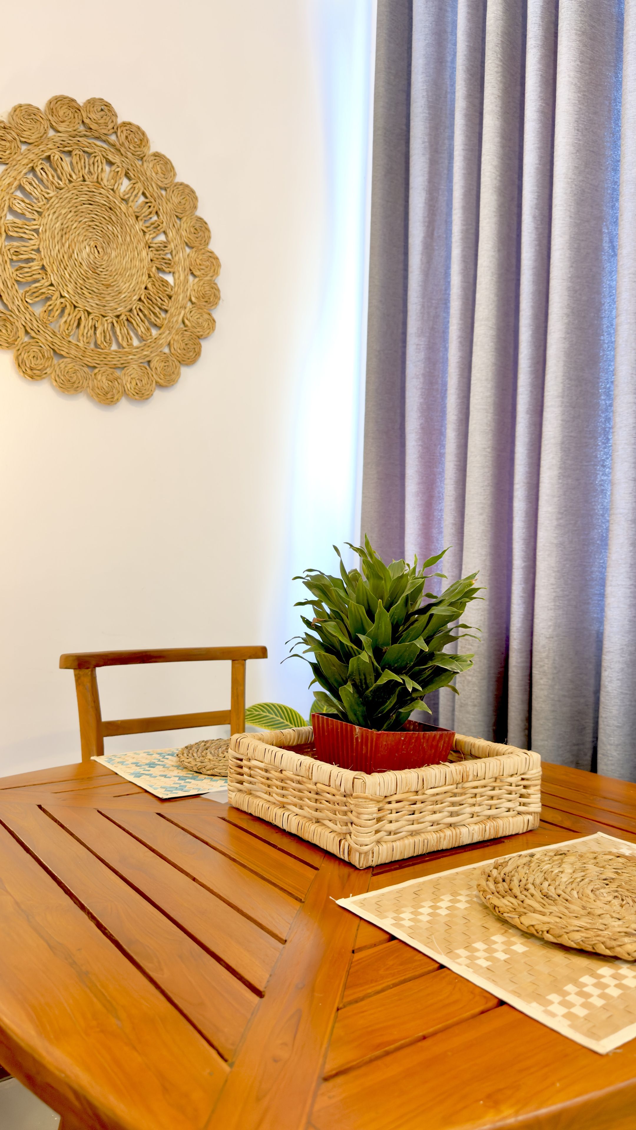 Une table de salle à manger en bois, surmontée d’un set de table en fibres tressées, un autre set de table en fibres tressées posé sur le sol, un panier tressé et une plante en pot.