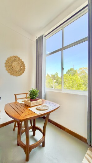 A room with a wooden table, chairs, a potted plant, a woven wall decoration, and a large window with curtains. - Crystal Pearl Villa Weligama (Weligama)