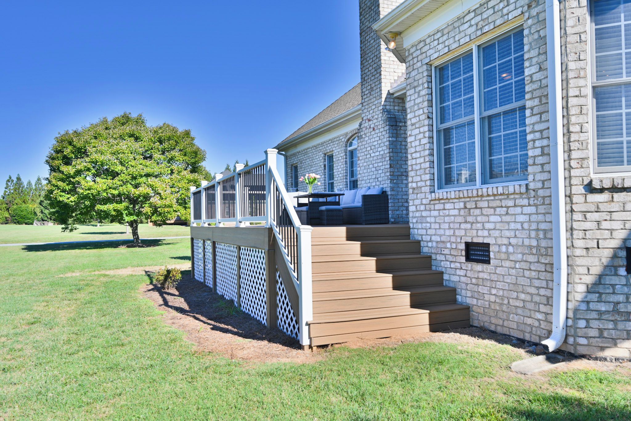 A house with a wooden deck, a staircase leading to a porch, and a large tree in the background.