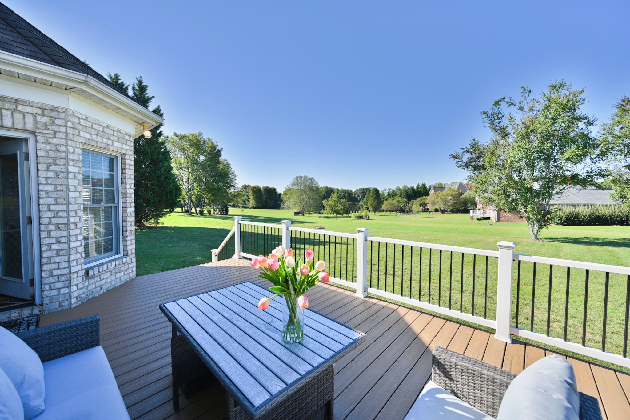 A deck with a table and chairs, a white railing, and a view of a green lawn and trees.