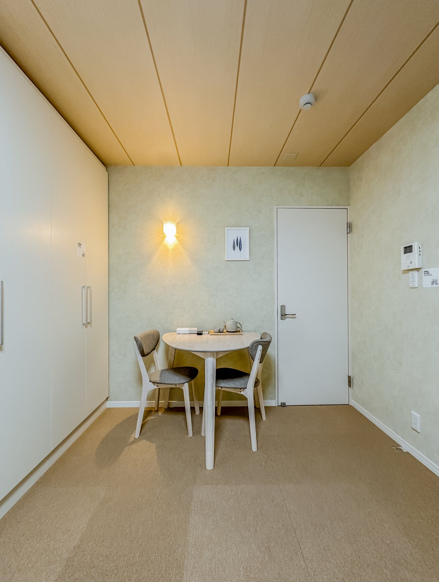 Una habitación pequeña y bien iluminada con una mesa redonda y dos sillas, una puerta y una lámpara fijada a la pared.