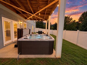 Jacuzzi en un patio trasero con terraza de madera, cerca viva blanca y vista a árboles y al atardecer.