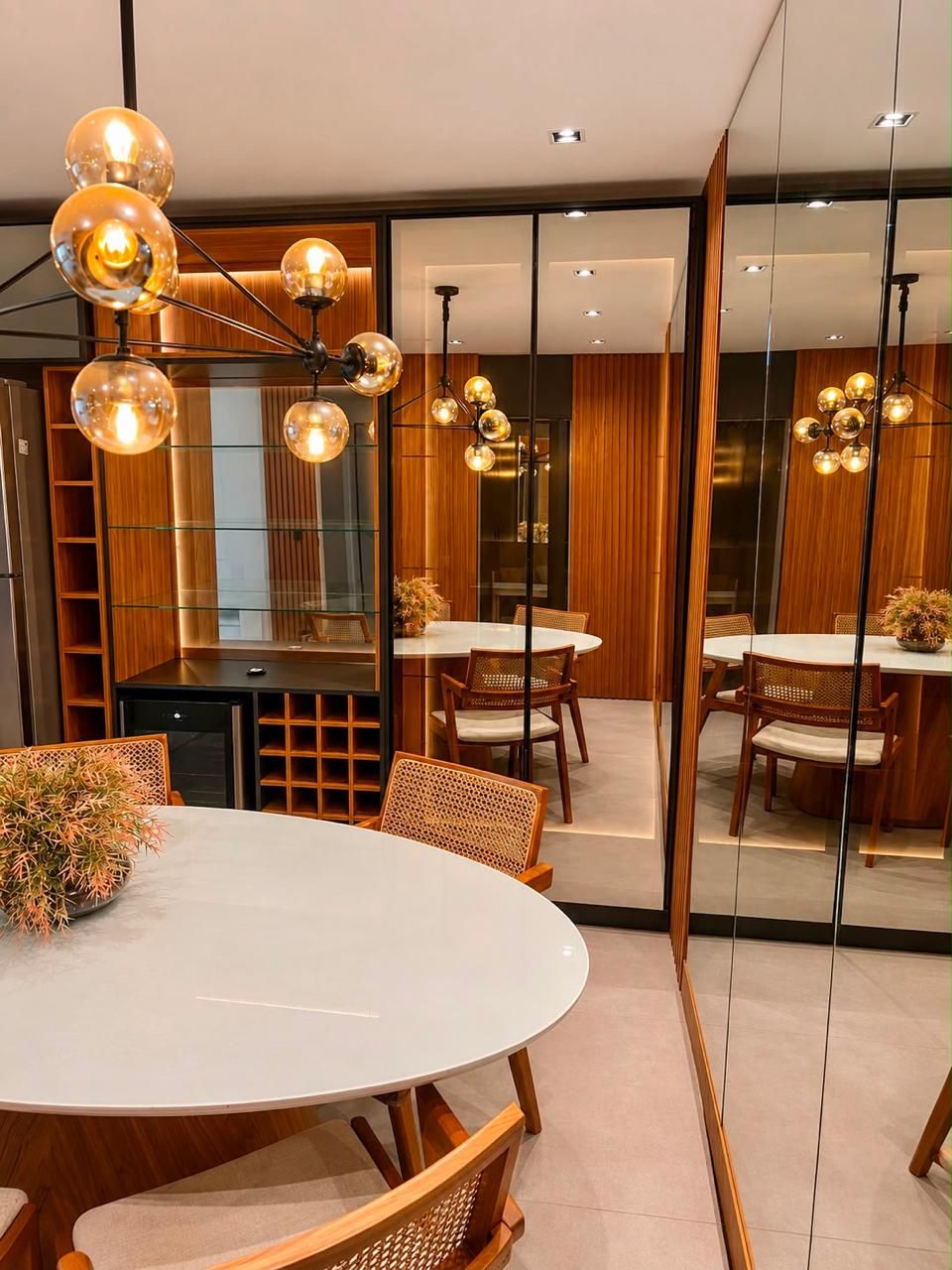 Un comedor moderno con mesas redondas, sillas de mimbre, una lámpara central y un panel de vidrio que refleja el ambiente.