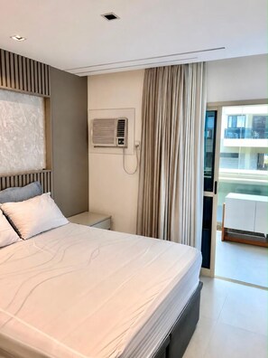 Ein modernes Hotelzimmer mit einem großen Bett, einem Nachttisch, einer eingebauten Klimaanlage und Blick ins Freie durch eine Glastür.