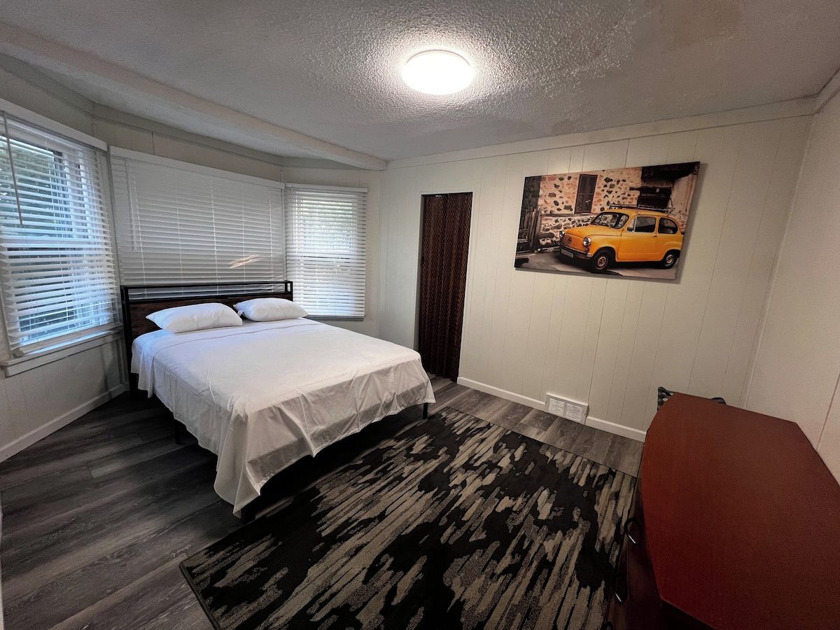 Un dormitorio con cama, una ventana con persianas, un cuadro colgado en la pared y una alfombra estampada.