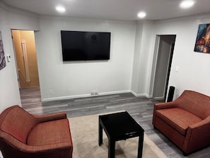Una habitación con una televisión de pantalla plana montada en la pared, dos sofás, una mesa de centro y suelo alfombrado.