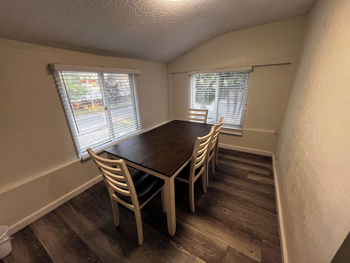 A dining room with a dark wooden table and four chairs, two windows with blinds, and wooden flooring.