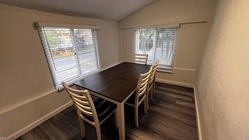 A dining room with a dark wooden table and four chairs, two windows with blinds, and wooden flooring.