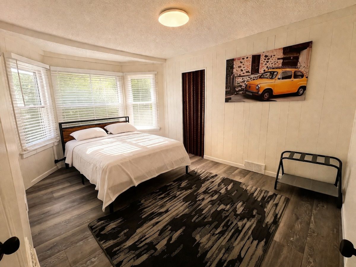 A bedroom with a bed, a window with blinds, a radiator, a rug, and a wall-mounted shelf.