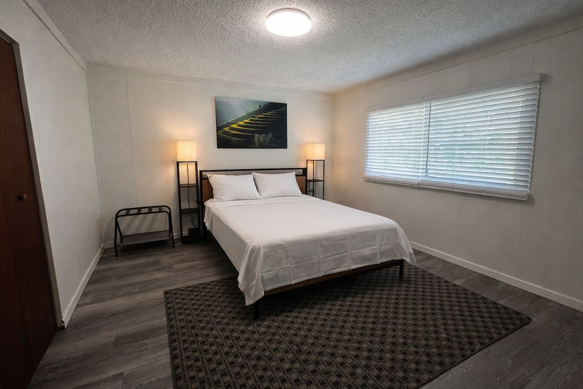A bedroom with a bed, bedside lamps, a window with blinds, a painting on the wall, and a patterned rug.
