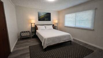 A bedroom with a bed, bedside lamps, a window with blinds, a painting on the wall, and a patterned rug.