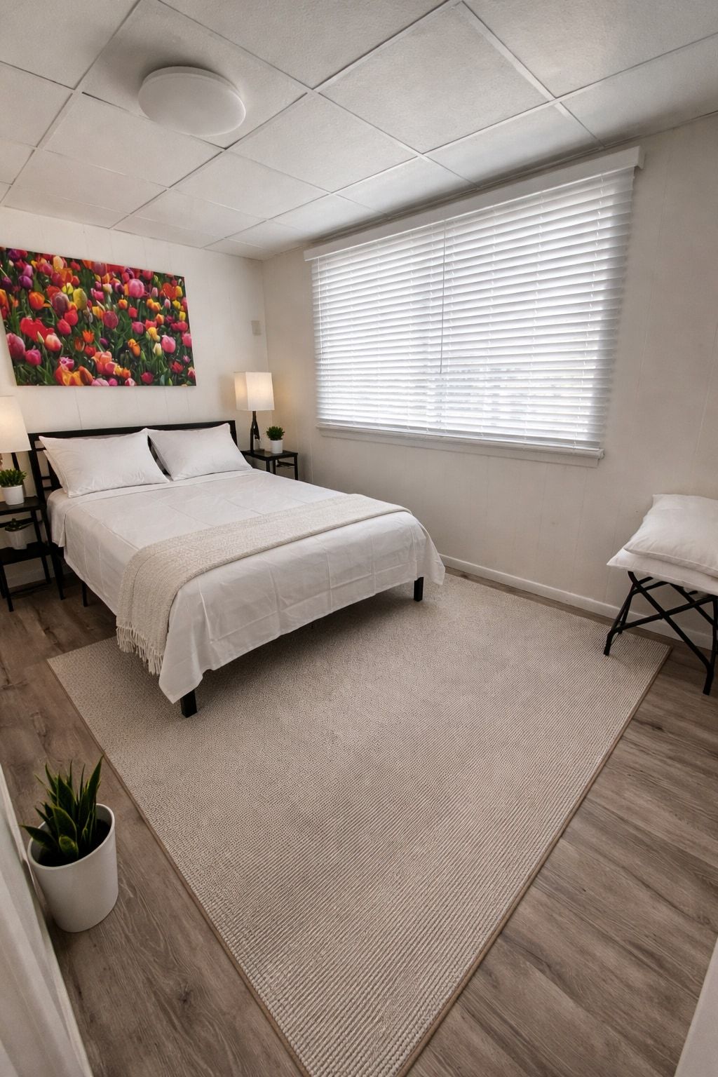A bedroom with a bed, bedside tables, a window with blinds, a painting on the wall, and a small table with a chair.