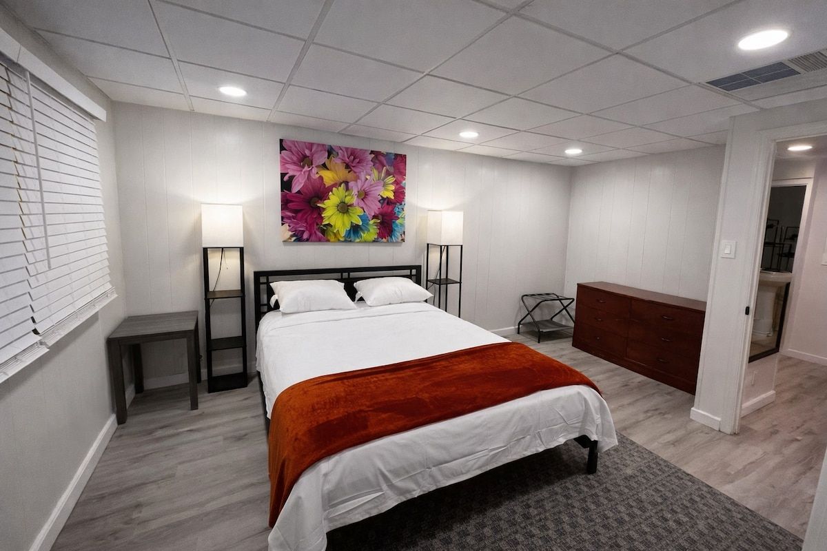 Quarto de hotel com cama, abajures, mesa de cabeceira, cômoda e uma grande obra de arte floral na parede.