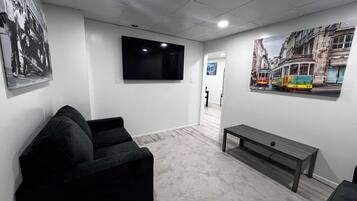 Une pièce avec un canapé noir, un banc en bois, un téléviseur à écran plat et un tableau représentant un paysage urbain accroché au mur.