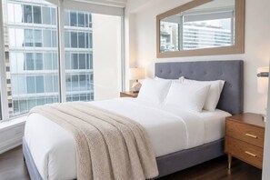 Ein Schlafzimmer mit einem großen Bett, einem Nachttisch, einem Spiegel und einem Fenster mit Blick auf Gebäude.