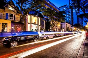 Eine belebte Stadtstraße bei Nacht mit modernen Gebäuden, einer Reihe geparkter Autos und einem Fußgängerweg.