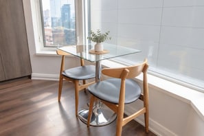 A modern dining area with a glass table and wooden chairs, a potted plant, and a city view through a large window. - Refined 1BR Suite-Workspace & Fast Wi-Fi (Toronto)