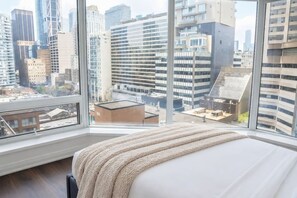 Habitació d'hotel amb un finestral gran que ofereix vista a la ciutat, un llit amb llençol beix i un disseny modern.