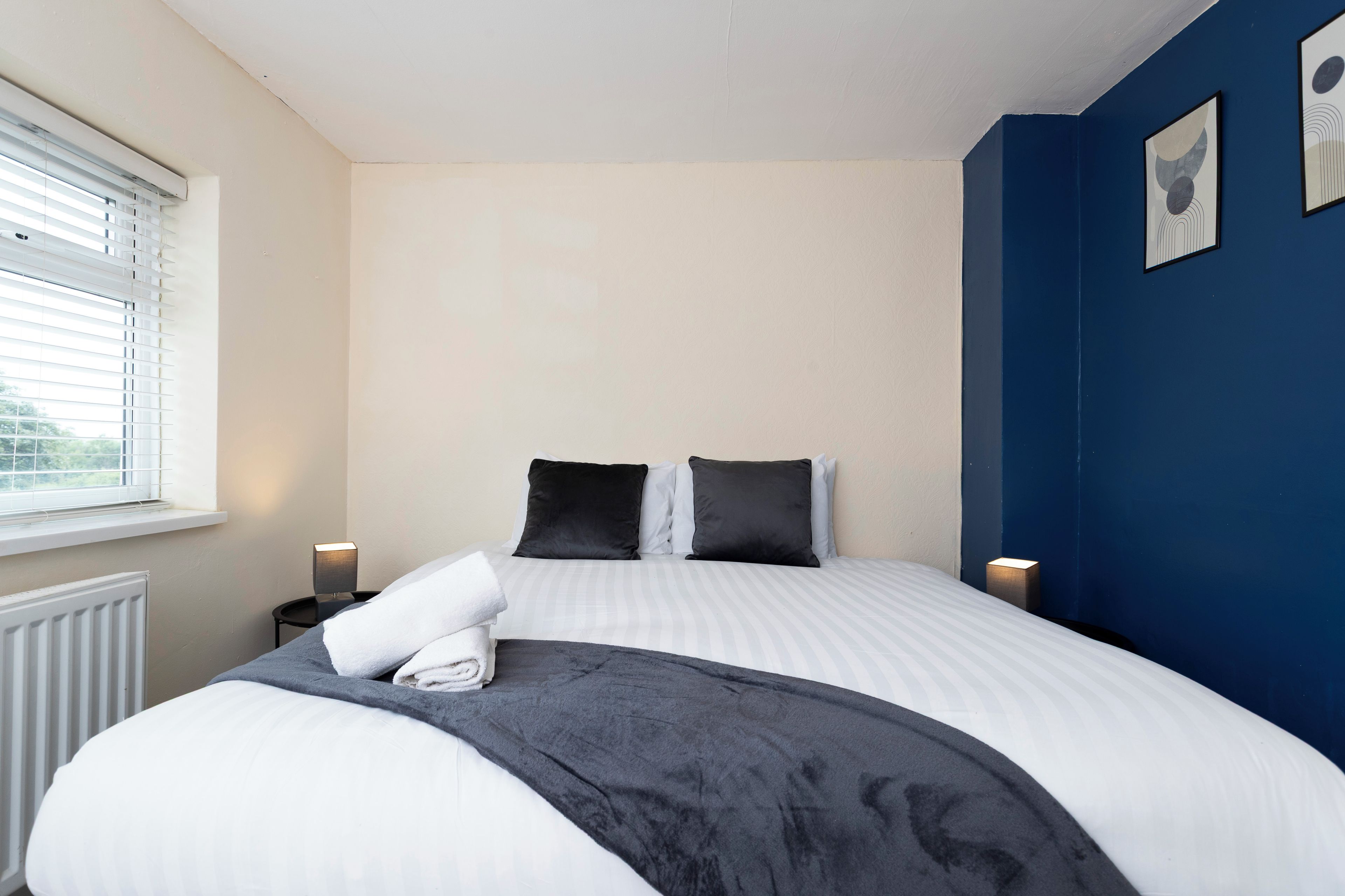 A bedroom with a bed, a window with blinds, a bedside table with a lamp, and a wall with a picture.