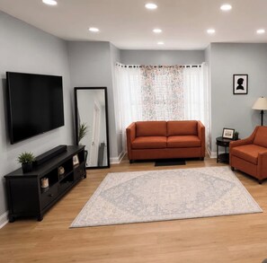 A living room with a large flat-screen TV, a sofa, a smaller chair, a rug, and a mirror. - Whole 621 Home - Sleeps 16 - Walk to Falls (Niagara Falls)