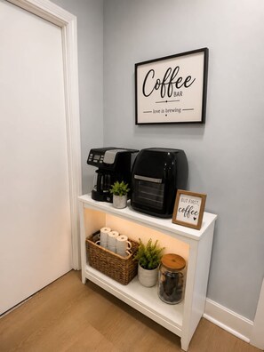 Ein Coffeeshop-Bereich mit einer Kaffeemaschine, einem Schild mit der Aufschrift 'Coffee Bar love is brewing' und einem gerahmten Schild, das 'But first, coffee' sagt.