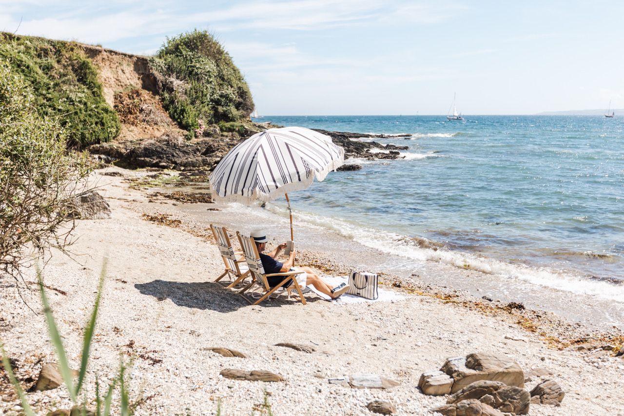 A beach scene with a person relaxing under a striped umbrella, a chair, and a bag on the sand.