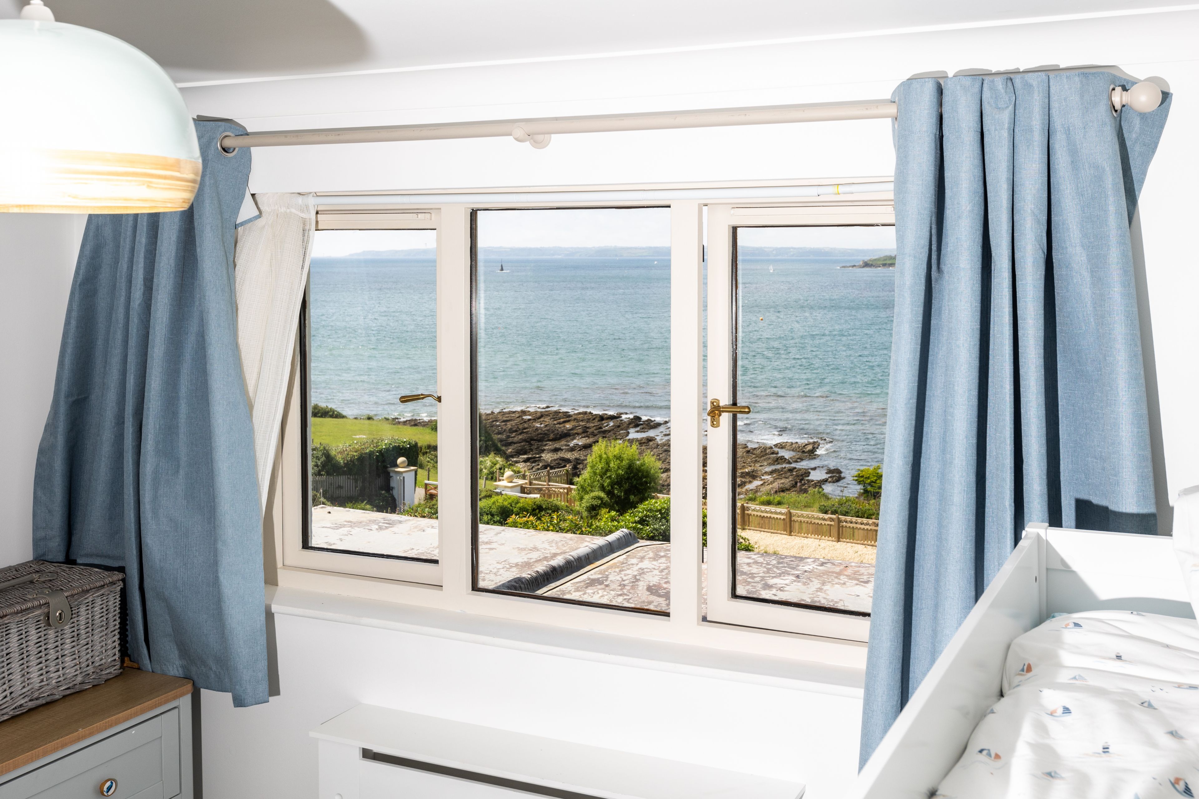 A bedroom with a view of the sea, a window with curtains, a bedside table, and a bed with a patterned bedspread.
