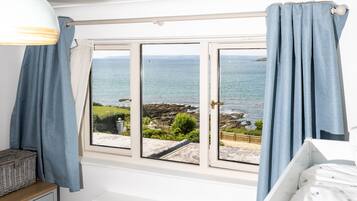 A bedroom with a view of the sea, a window with curtains, a bedside table, and a bed with a patterned bedspread.