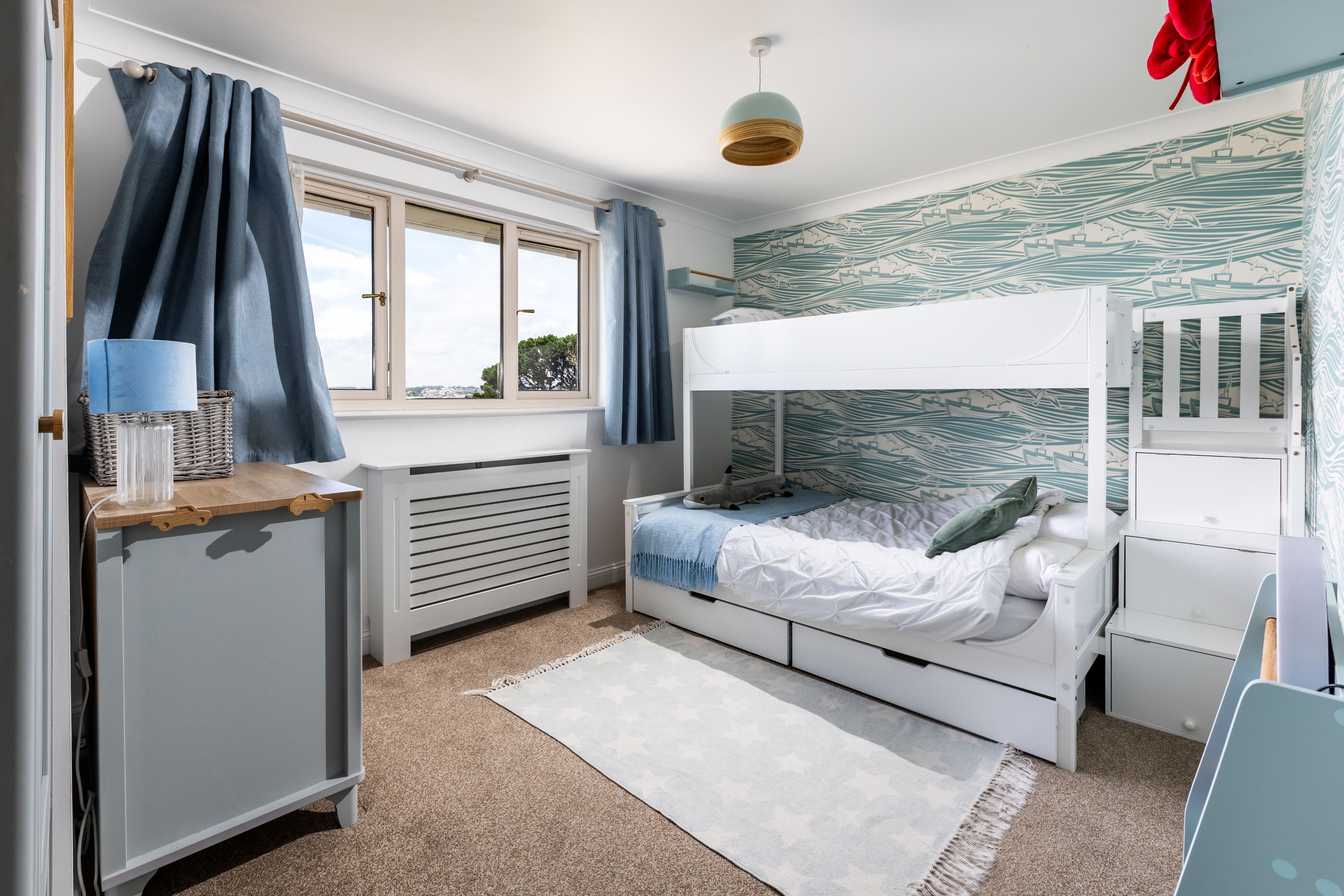 Una cama individual superpuesta con un mural de temática náutica, un escritorio con una lámpara y una ventana con cortinas.
