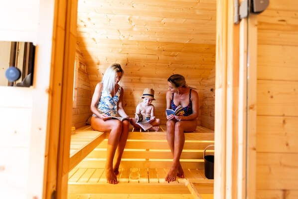 Drei Personen sitzen in einer Holzsauna mit Bänken, Lektüre und einem kleinen schwarzen Behälter auf dem Boden.
