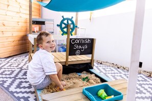 Lapsi leikkii hiekkalaatikossa, jossa on kyltti, jossa lukee: ”Cornish pysyy” sekä laivan ruorin kuva.