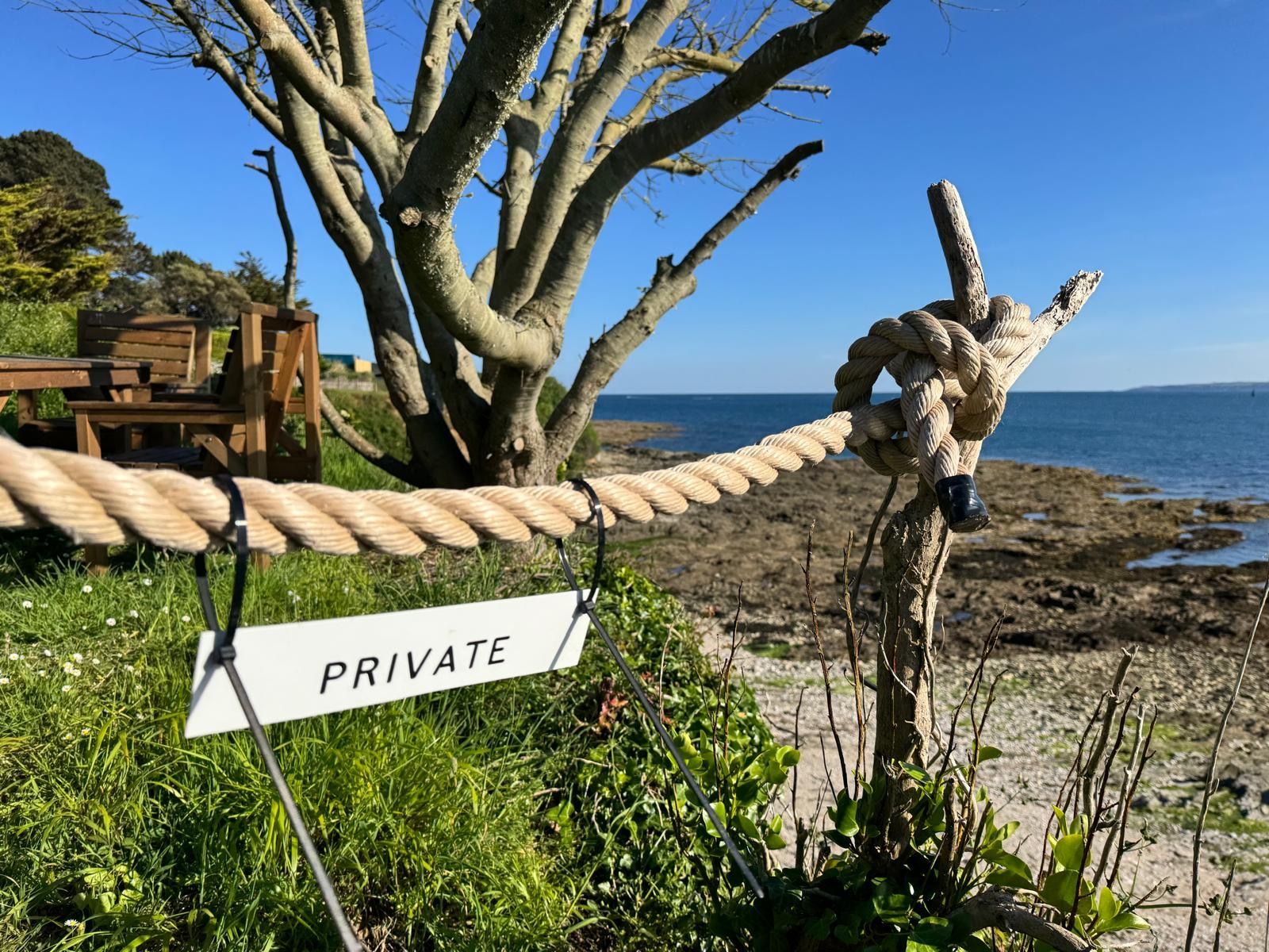 A rope barrier with a 'PRIVATE' sign, a wooden bench, and a tree by a beach.
