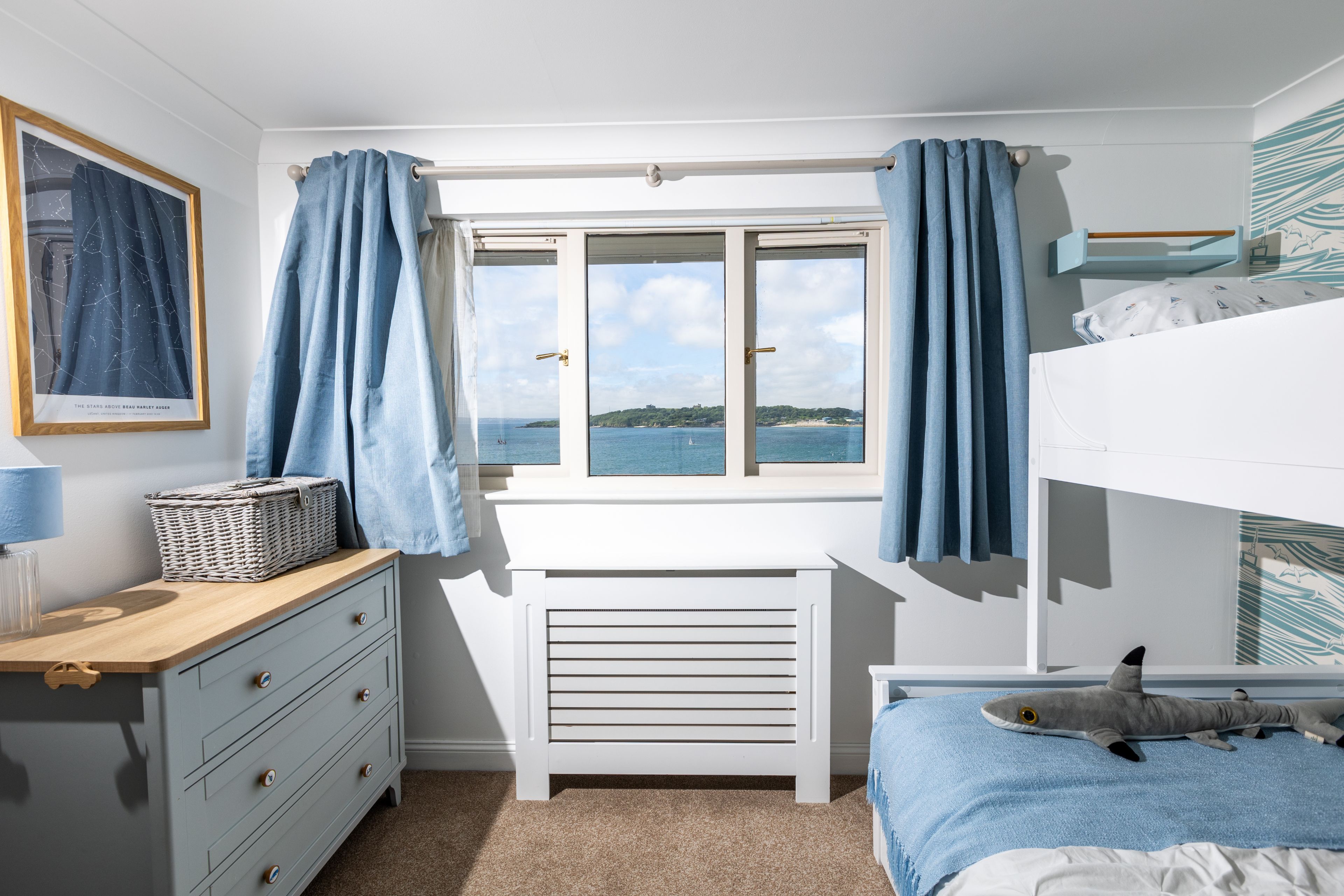 A bedroom with a view of the sea, a bunk bed, a desk with a lamp, and a dresser.